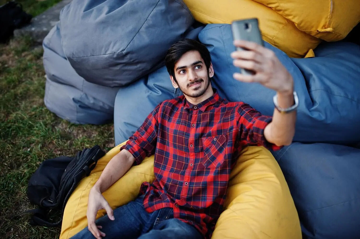 Junger indischer Student im karierten Hemd und in Jeans sitzt auf Outdoor-Kissen, entspannt sich und macht ein Selfie.
